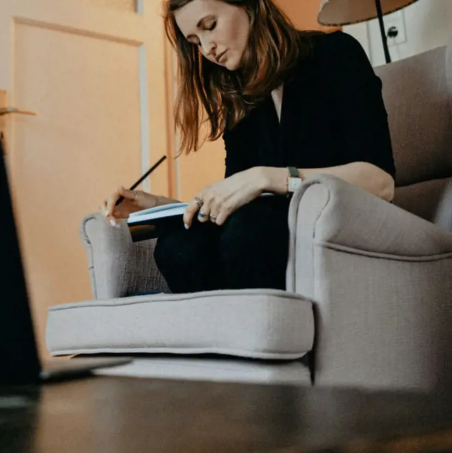 Young woman in black sits in a gray armchair, writing in a notebook at home; warm light and quiet surroundings create a thoughtful mood.