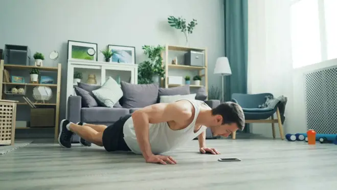 Ein Mann in Tanktop und Shorts macht einen Liegestütz in einem hellen Wohnzimmer. Die Umgebung mit Sofa, Regalen mit Pflanzen und einer Trainingsmatte vermittelt Fokus und Entschlossenheit.