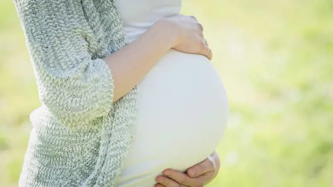 Pregnant person in a soft gray knit sweater cradles their belly outdoors, conveying tenderness, comfort, and joyful anticipation.