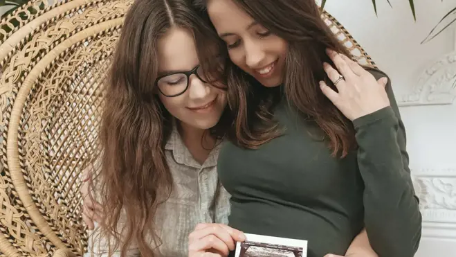Zwei Frauen umarmen sich in einem Korbsessel, lächeln und halten ein Ultraschallbild an einen schwangeren Bauch in einem gemütlichen Zuhause mit Pflanzen.