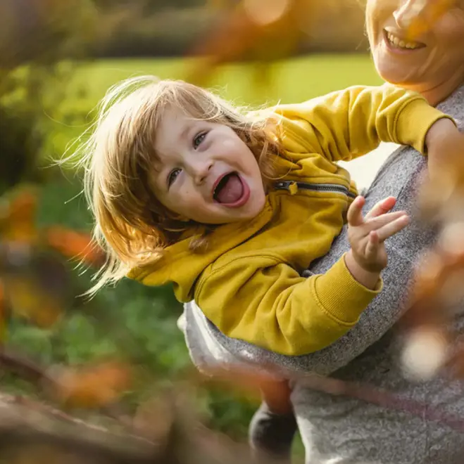 Smiling toddler in yellow hoodie laughs while being held by a woman outdoors in a sunny park with blurred autumn leaves.