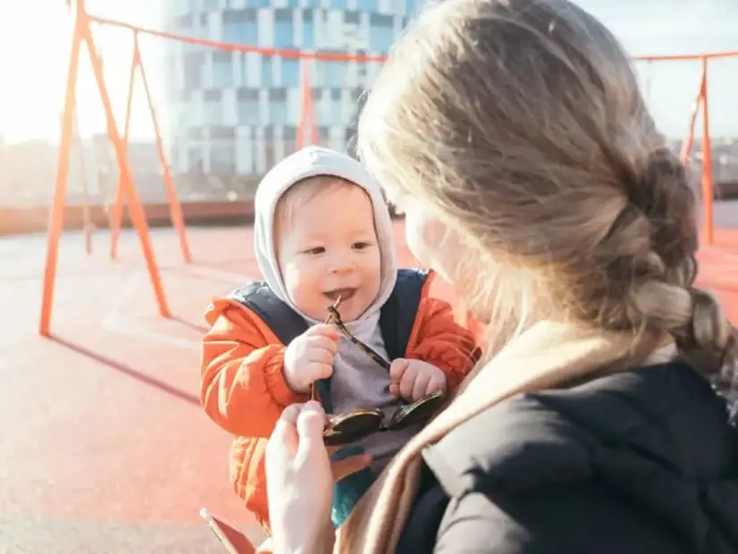 Mum holds sunglasses as baby in orange jacket and grey hood chews the arm, smiling at her on a sunlit playground with swings and a modern building behind.