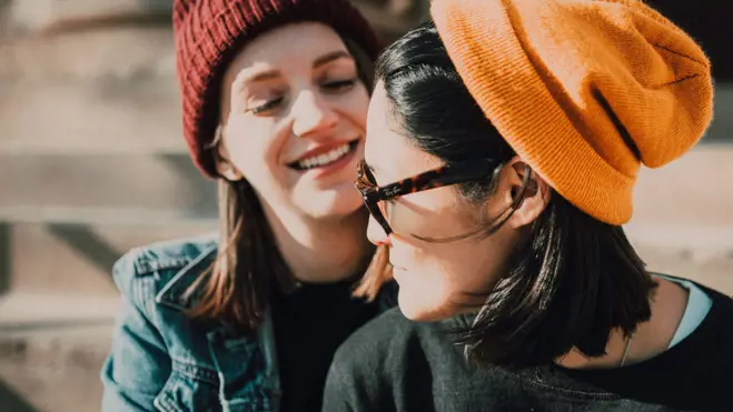 Two smiling women in knit beanies share a joyful outdoor moment; one wears sunglasses and an orange hat, the other a denim jacket.