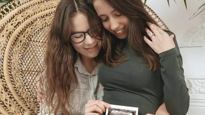 Two women embrace in a wicker chair, smiling while holding an ultrasound scan photo against a pregnant belly in a cosy home with plants.