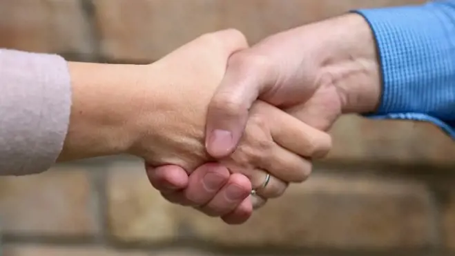 Ein Mann und eine Frau geben sich vor einer strukturierten Backsteinwand die Hand, was Einigung oder Partnerschaft symbolisiert
