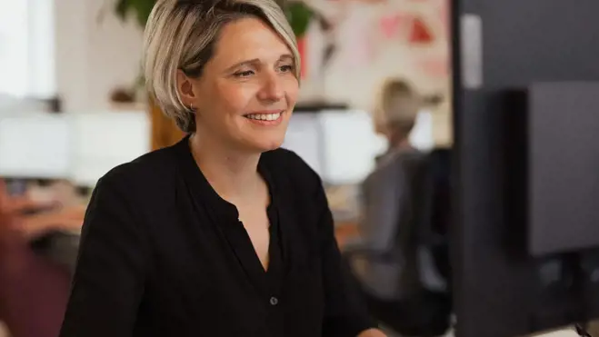 Smiling woman in a black blouse works at a desktop computer in a bright, modern office, conveying confidence, focus, and a positive mood.