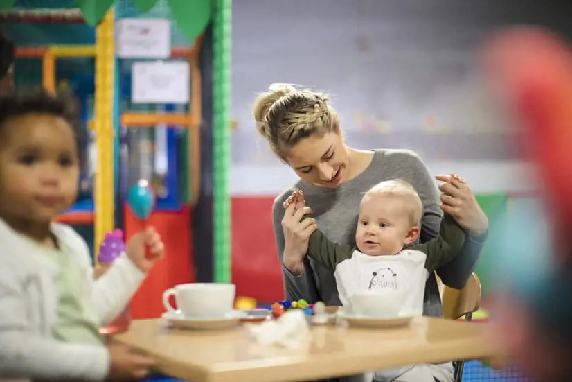 Woman holds baby’s hands at a nursery table while a toddler watches, with cups and small toys in a colourful indoor play area.