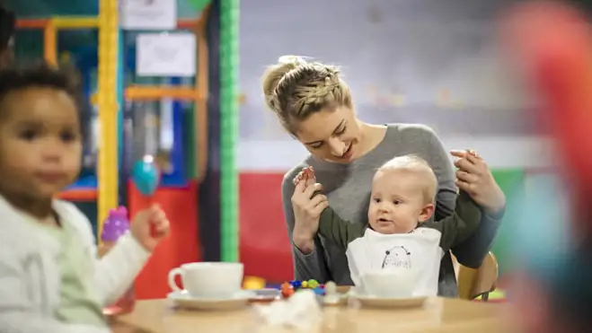 Woman holds baby’s hands at a nursery table while a toddler watches, with cups and small toys in a colourful indoor play area.