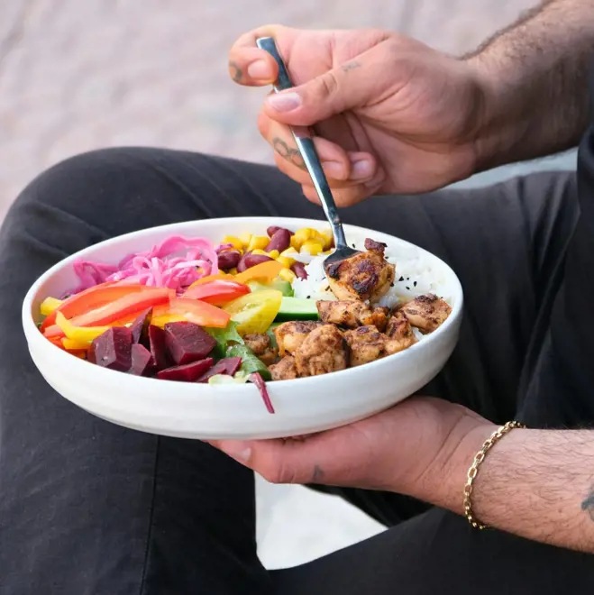 A person holds a bowl of vibrant food, with grilled chicken, fresh vegetables, rice, and beans. The setting is casual, conveying a relaxed mood.