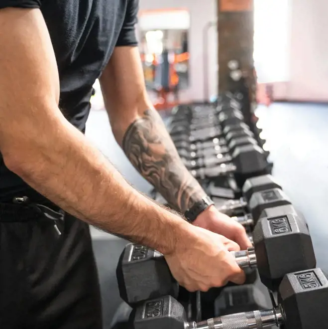 Eine Person mit tätowiertem Arm nimmt in einem Fitnessstudio eine Hantel aus einem Hantelständer. Die Umgebung vermittelt Konzentration und Fitness.