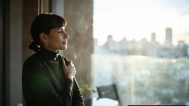 Woman in a black turtleneck stands by a window, gazing at a sunlit city skyline, with balcony furniture and reflections on the glass in an apartment.
