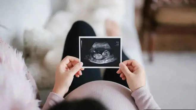 Person hält ein Ultraschallbild über einem schwangeren Bauch, während sie zu Hause auf einem Sofa sitzt.