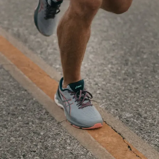Läufer in grauen Turnschuhen joggt auf einer Straße, der Fokus liegt auf den Beinen. Die Szene vermittelt Bewegung und Entschlossenheit, mit einer sichtbaren gelben Linie.