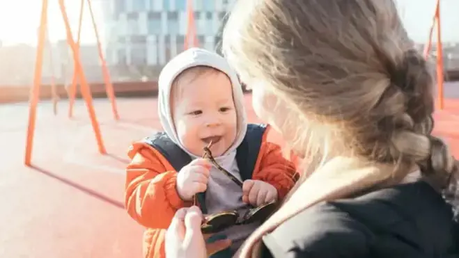 Eine Mutter hält eine Sonnenbrille, während ein Baby in einer orangefarbenen Jacke und grauen Kapuze an einem Bügel kaut und sie auf einem sonnigen Spielplatz mit Schaukeln und einem modernen Gebäude im Hintergrund anlächelt.