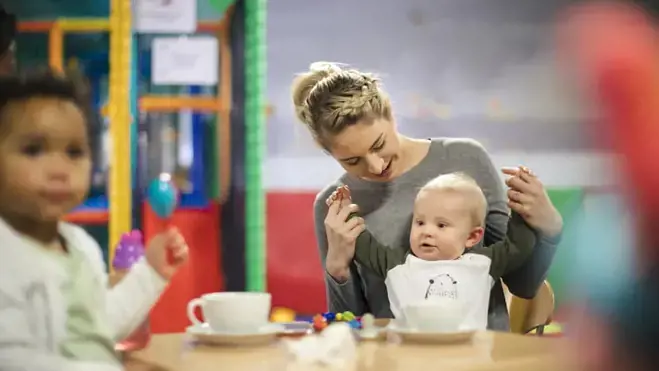 Eine Frau hält die Hände eines Babys an einem Tisch in einer Kindertagesstätte, während ein Kleinkind zusieht; Becher und kleine Spielzeuge stehen in einem farbenfrohen Indoor-Spielbereich.